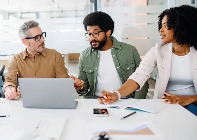 Three diverse professionals engaged in a discussion at a modern office table, with a laptop and documents in front of them.