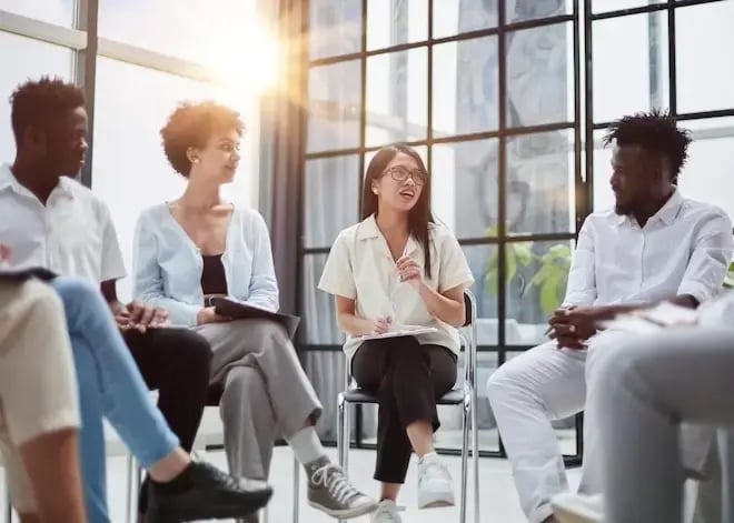 A diverse group of professionals sits in a bright room, engaged in a focus group discussion, with sunlight streaming in.