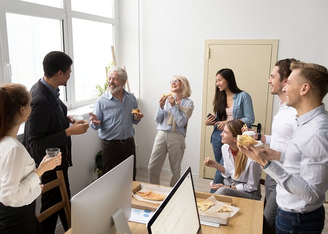 A diverse group of eight people of various ages, smiling and laughing, gather around a desk in a bright office space, eating pizza and holding drinks, suggesting a casual and convivial atmosphere.