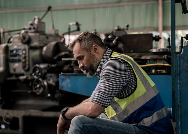 A tired factory worker in a reflective vest sits on a bench, looking down with a weary expression in an industrial setting.