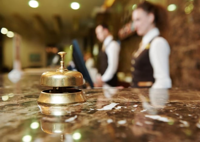 A polished brass service bell sits on a reflective marble hotel reception desk. Two blurred hotel staff members in vests and white shirts stand in the background, behind the counter. Soft, warm lighting illuminates the scene.