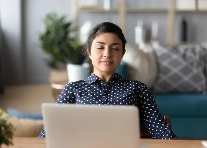 A woman in a polka dot blouse sits at her desk with eyes closed, looking calm and focused, with a laptop in front of her.