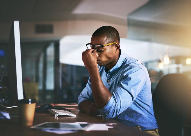 A man in glasses and a blue shirt sits at his desk, rubbing his forehead in frustration, with a computer screen in front of him.