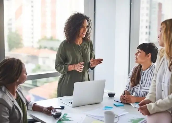 A confident woman leads a team meeting in a modern office, speaking to colleagues seated around a table with laptops and notes.