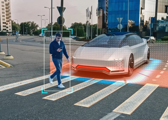 A silver autonomous vehicle is shown at a crosswalk in a city. The car appears to be detecting a person, looking at their phone while walking across the pedestrian crossing.