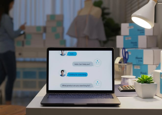 A laptop on a desk displays a customer service chatbot interface. The background shows a person packing boxes in what appears to be a small business or home office setting with shipping supplies, suggesting the chatbot is for an e-commerce business.