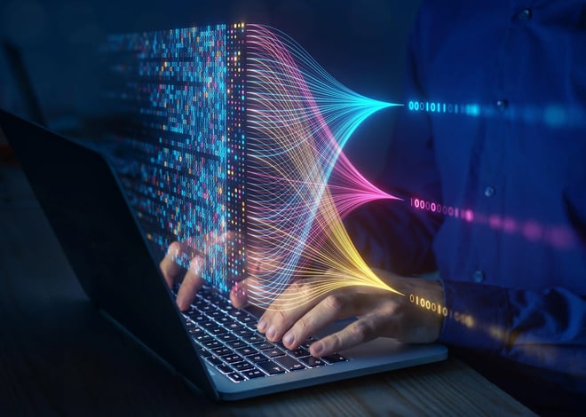 A person's hands are shown typing on a laptop keyboard. Emerging from the laptop screen is a vibrant, abstract data visualization resembling a cube of digital information with colorful streams flowing outward, converging into binary code representations.