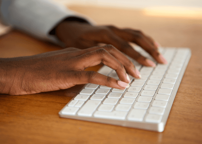 Close-up of hands typing on a wireless keyboard on a wooden desk.