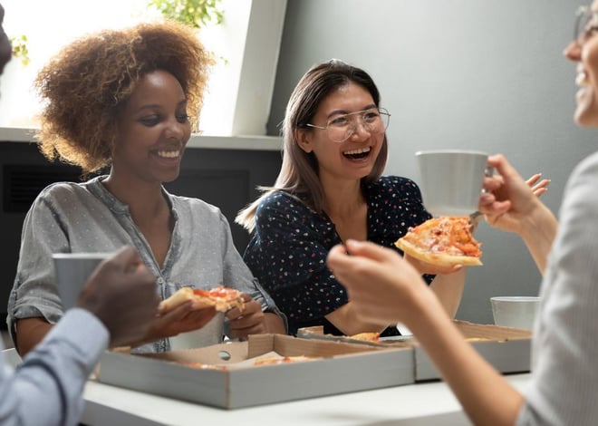 Four diverse colleagues are gathered around a table, joyfully eating pizza and holding mugs. Two pizza boxes sit on the table, with slices remaining. The atmosphere appears relaxed and convivial, suggesting a team lunch or casual gathering.