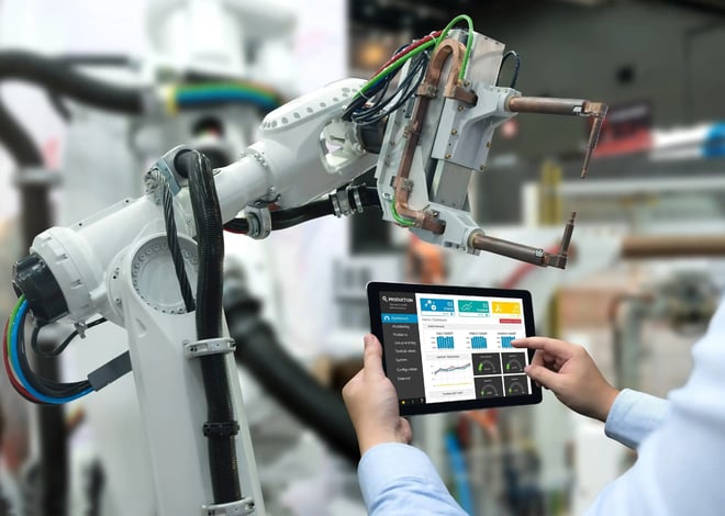 A person holds a tablet displaying a control interface with charts and data, facing a large white industrial robot arm in a factory setting.