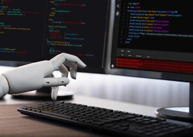 A robotic white hand hovers over a black computer keyboard in front of two monitors displaying lines of colorful code on a dark background.
