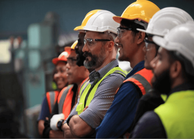 A group of workers wearing hard hats and safety vests standing together in an industrial setting. They appear to be listening attentively, with one individual in focus wearing a white helmet and a reflective vest.