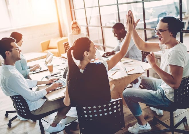 A diverse group of six colleagues are gathered around a light wooden table in a bright, modern office space. Two individuals in the foreground, a woman with a high ponytail and a man with glasses and a beanie, are enthusiastically giving each other a high-five, expressing excitement and success.