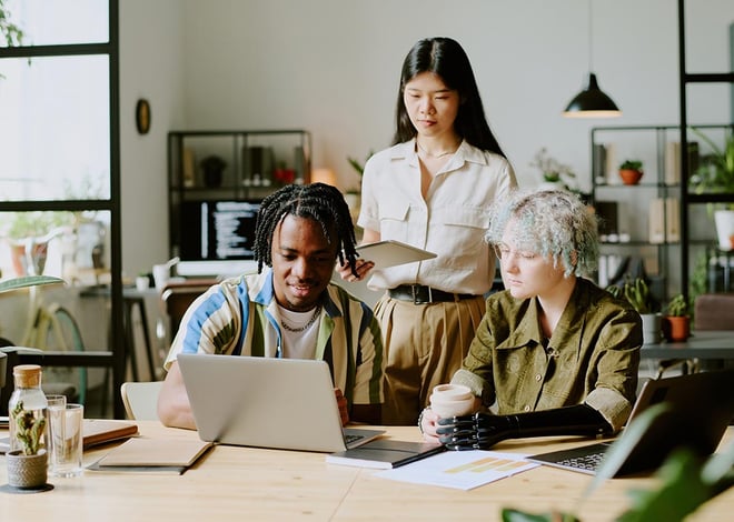 Three young, diverse colleagues collaborating at a wooden table in a modern, sunlit office. A Black man is smiling and pointing at his laptop screen. An Asian woman stands behind him, holding a tablet.
