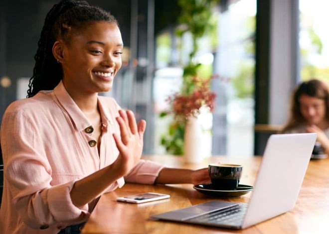 A person smiling and waving at a laptop screen, seemingly at a coworking space.