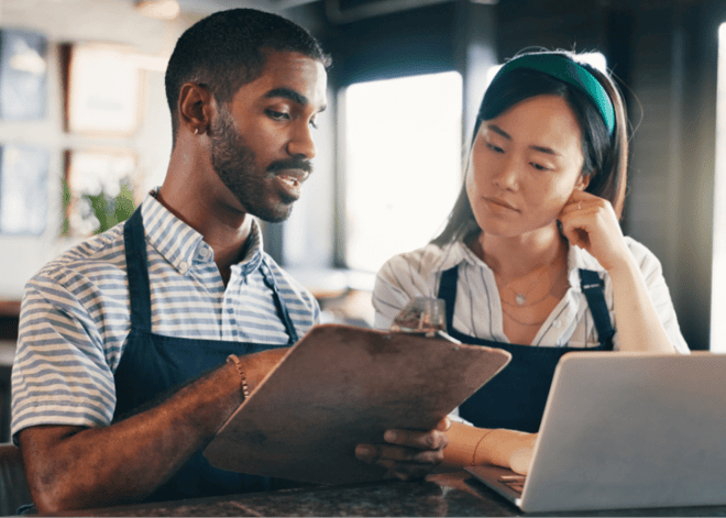 A man holding a clipboard discusses work with a woman using a laptop, both wearing aprons in a professional setting.