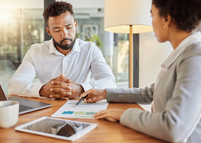 A serious discussion between a man and a woman at a desk with documents, a tablet, and a laptop.